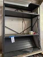 Black metal shelving unit with four shelves total, one shelf detached, shown in garage with items on shelf and black panel below