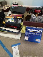 Various office supplies piled on the floor including binders, folders, papers, and an Olympus Stylus 400 Digital camera box.