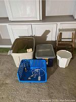 Photo of a large beige storage tub, a gray lidded file tote, a blue basket with cords, a white trash bin and a brown wooden step stool.
