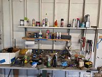 View of garage workbench surface showing assorted cans, bottles of chemicals and lubricants, old license plates on shelves, and miscellaneous small tools and hardware items.