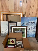 Photo showing assortment of framed art and two large empty frames. Art includes nautical, car, and winter landscape themes. Box with several small frames contains artwork with hearts and nature.