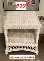 Front view of white wicker side table showing woven top surface and lower shelf with braided trim edges, photographed against a wall with framed posters.