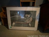 Front view of framed print showing person with furry hood, visible broken glass on right side, signed illegibly on bottom right corner.