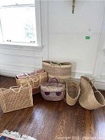 Six woven beach bags arranged on a hardwood floor showing variety in size, shape, material texture, and handle color.