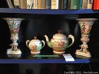 Front view of the tea pot, sugar bowl and two matching vases on a shelf, showing detailed painted scenes and ornate bases.
