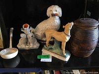 Overview of shelf showing white pipe-bearing dog figurine, tan greyhound, double poodle vase, ceramic ladle and wooden container