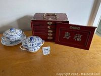 Overall view of red wood jewelry box with detached panel, two lidded porcelain bowls, and saucer on table