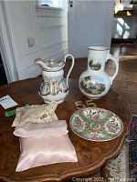 Overview of two porcelain jugs, decorative plate, two pillows, and gold toned pins displayed on a wooden table with natural lighting.