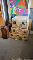 Four vintage luggage pieces including three cream-colored trunks stacked and one brown suitcase leaning against the wall, showing overall look and stacking.