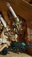 Two vintage sconces, a brass one with two candle-shaped light holders and a black cast iron one, on packaging paper inside a cardboard box.