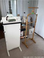 Photo showing white Corny of Windsor trouser press and wooden clothes butler upright on carpeted surface near white doors and cardboard boxes.
