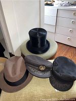 Four men’s hats (top hat, fedora, two caps) displayed on ottoman in front of dresser