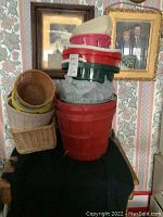 Stack of assorted plastic pails in red, green, white, beige, one tin pail, and wicker baskets on side