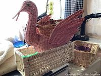 Large pink swan-shaped woven basket with wings extended sitting inside a natural rectangular basket, shown with other baskets in background