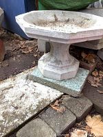 Side view showing the full bird bath structure with a hexagonal pedestal base set on stone slabs and leaves on the ground.
