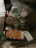 Photo of various canning supplies including glass jars, metal lids, screw bands, box of accessories, and a jar opener tool viewed from the top