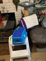 Photo showing the cat play stand with multiple levels and sisal-wrapped scratching posts, three stacked plastic stools (two white and one blue), and a white fleece cat blanket with purple border draped on the stand.