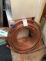 Full coil of copper tubing placed indoors on plywood floor, showing entire coil and tagged with 'copper' label.