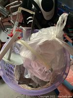 Plastic lavender-colored basket containing multiple metal doll stands and other craft items inside a plastic bag.