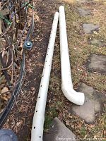 Two white PVC downspout pipes on grass and stone path, one pipe with multiple round holes, other pipe with elbow fitting.