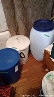 Photo showing all three items: white metal garbage can with black handles and lid, blue plastic bucket with lid, and large white plastic container with blue lid.