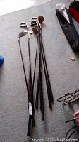 Photo showing eight golf clubs laid out on carpet floor, including multiple irons, drivers, wood heads, and putter with grips visible.