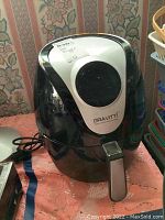 Front view of Gravitti air fryer with power cord visible, sitting on patterned cloth surface.