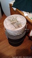 Two round hat boxes stacked; top box has a floral print with plants in pots; bottom box is dark and plain.