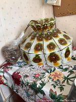 Front angle showing the full lamp with colorful textured glass floral panels and scalloped top edge. Lamp is placed on table with visible cord and wooden block hanger.