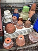 Photo showing a variety of terra cotta clay pots with white patina, green and blue resin planters, a white ornate ceramic base, and tall orange clay planters arranged on outdoor surface.