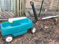 Image showing the entire blue plastic Step 2 wagon with a black pull handle and white wheels, placed outdoors on a dirt surface.