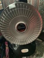 Front view of Presto HeatDish electric heater showing wire mesh reflector and central logo, with black base and control knob visible.
