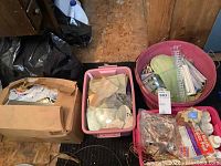 Overview photo of four bins with stained glass pieces, wire coils, cleaning chemicals and lamp supplies in plastic and cardboard bins on wooden floor in basement.