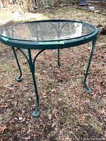 Outdoor round side table with metal frame and clear removable glass top. The table has a green metal rim and curved legs with circular foot pads. Visible scratches on the metal frame.