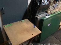 Wood folding side table with natural finish showing top and folded legs, placed on a rug next to other tables.