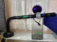 Image showing adjustable black task light with tape covering, large rectangular glass bottle, and purple glass ball with cork stopper on top of the bottle against a lace curtain background.