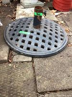 Cast iron umbrella stand showing lattice patterned base and umbrella holder tube with knob, placed outdoors on a stone surface with leaves around.
