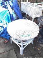 Round white wicker table with woven top and slatted lower shelf, showing outdoor location and some surface wear.