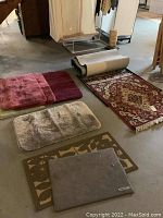 Collection of various rugs including small bathroom rugs, area rug, doormats, and rug remnants laid out on basement floor.