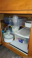 Photo of lower cabinet shelf showing plastic storage containers, small pitcher, white ceramic casserole dish, and measuring utensils