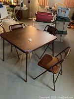 Wide view of the square folding card table with 3 matching folding chairs in a basement with storage bins and other household items.