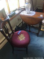 Wooden chair with carved backrest and red floral upholstered seat placed by window next to drop leaf side table holding a crystal vase with doily