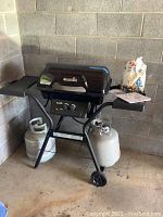 Full view of the Char-Broil black propane grill with two propane tanks and a bag of hickory chips placed on the side tray.