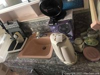 Bathroom countertop with a pink sink, small black Holmes desk fan with box, white analog scale, glass jars with lids, and a cream fabric shaver.