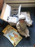 Photo of Shirley Temple collector doll in cream jumper and hat seated next to 3 vintage Shirley Temple paper dolls books on a blue carpet.