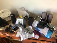 Photo showing assorted electronics on wooden table including Sirius satellite radio, Dell speakers, Omron BP monitor, Jitterbug phone, Nikon camera, various cables and manuals.