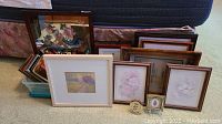 Photo of all the framed art pieces and picture frames, stacked and leaning on each other, with a plastic basket visible at bottom left corner. Various sizes and frame types visible.