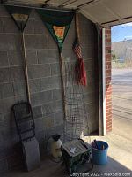 Two garden rakes hanging on wall, three wire tomato towers on the floor, plastic sprayer, garden stool, and bucket with hand tools
