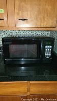 Front view of black Hamilton Beach microwave oven on kitchen counter with cabinets and backsplash.