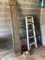 Painted plank, wooden ladder, adjustable hand truck, and traction kit box beside a climate concrete block wall with garden tools hanging nearby.